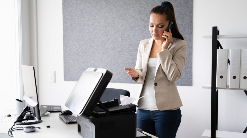 A woman is on her phone looking at a printer that has a paper jam.