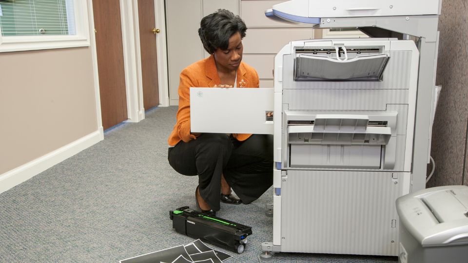 Frustrated business woman fixes copier, works on replacing toner cartridge and papers are scattered on the floor.