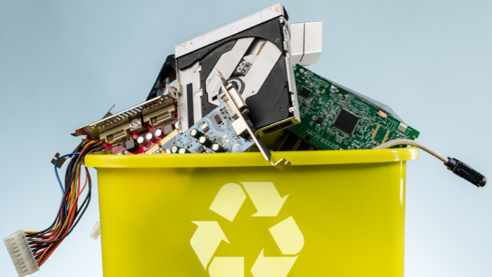 A green trash bin with a recycling logo is filled with electronic items.